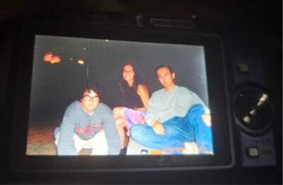 A photo of a camera displaying a picture of three young adults. The girl in the middle is smiling, the boys around her are staring into the camera